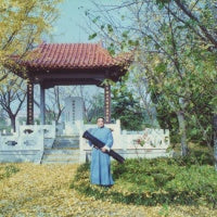 Master Mei Riqiang holding his guqin in a classical Chinese garden, wearing a traditional blue robe, autumn leaves on the ground
