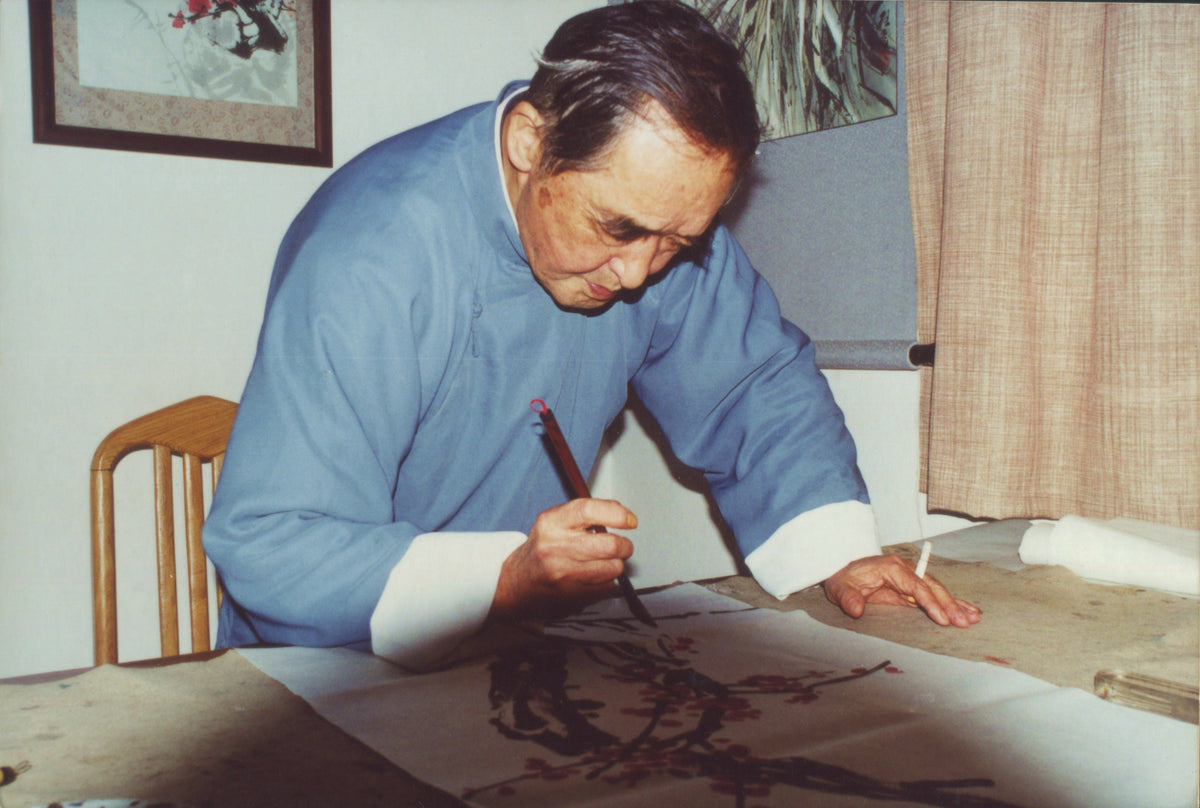 Master Mei Yueqiang painting plum blossoms at his desk, brush in hand, wearing a blue scholar's robe