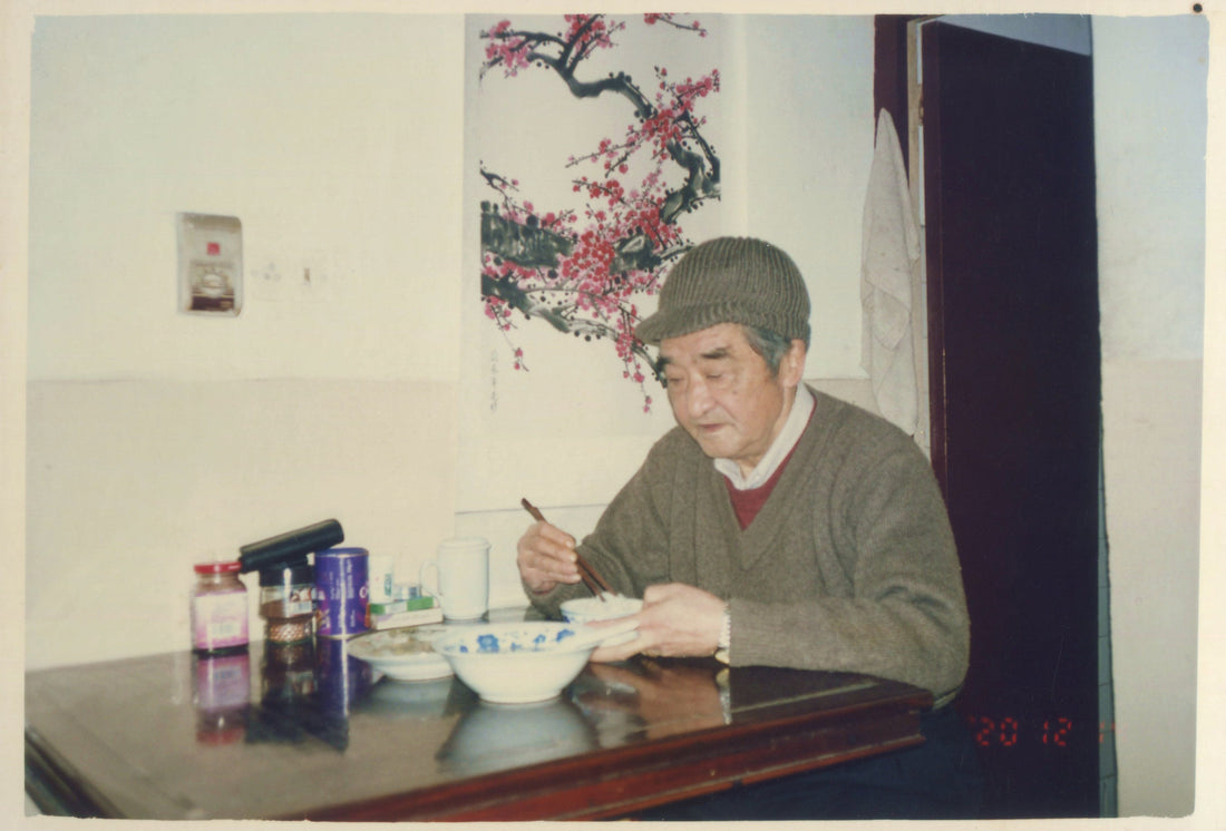 Master Mei Yueqiang eating at his small table in Jiandao Lane, Yangzhou, a plum blossom painting on the wall behind him — savoring, not craving