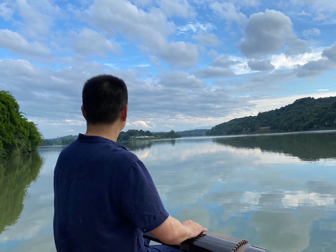 A man sits by a tranquil lake playing the guqin, viewed from behind, with clouds reflected on the still water